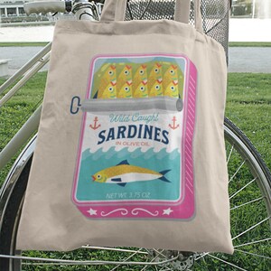 May include: A beige canvas tote bag with a pink and blue illustration of a can of sardines. The can has the text "Wild Caught Sardines in Olive Oil" and "Net Wt. 3.75 oz". The bag is hanging on a bicycle.