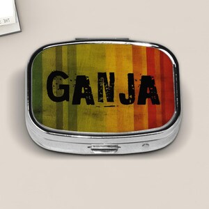 May include: A silver pill box with a colorful design. The top features the word "GANJA" in bold black letters over a background of vertical stripes in green, yellow, and red.