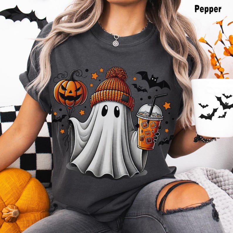 Puede incluir: Una camiseta gris con un fantasma de dibujos animados que lleva un gorro de punto y sostiene una calabaza y una taza de caf&eacute; helado. El fantasma est&aacute; rodeado de murci&eacute;lagos y estrellas.