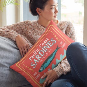 May include: Red decorative pillow with a vintage-style sardine can label design. The label reads "Pacific Coast Sardines in Olive Oil" with a blue fish illustration and text in a yellow border. The pillow is rectangular.