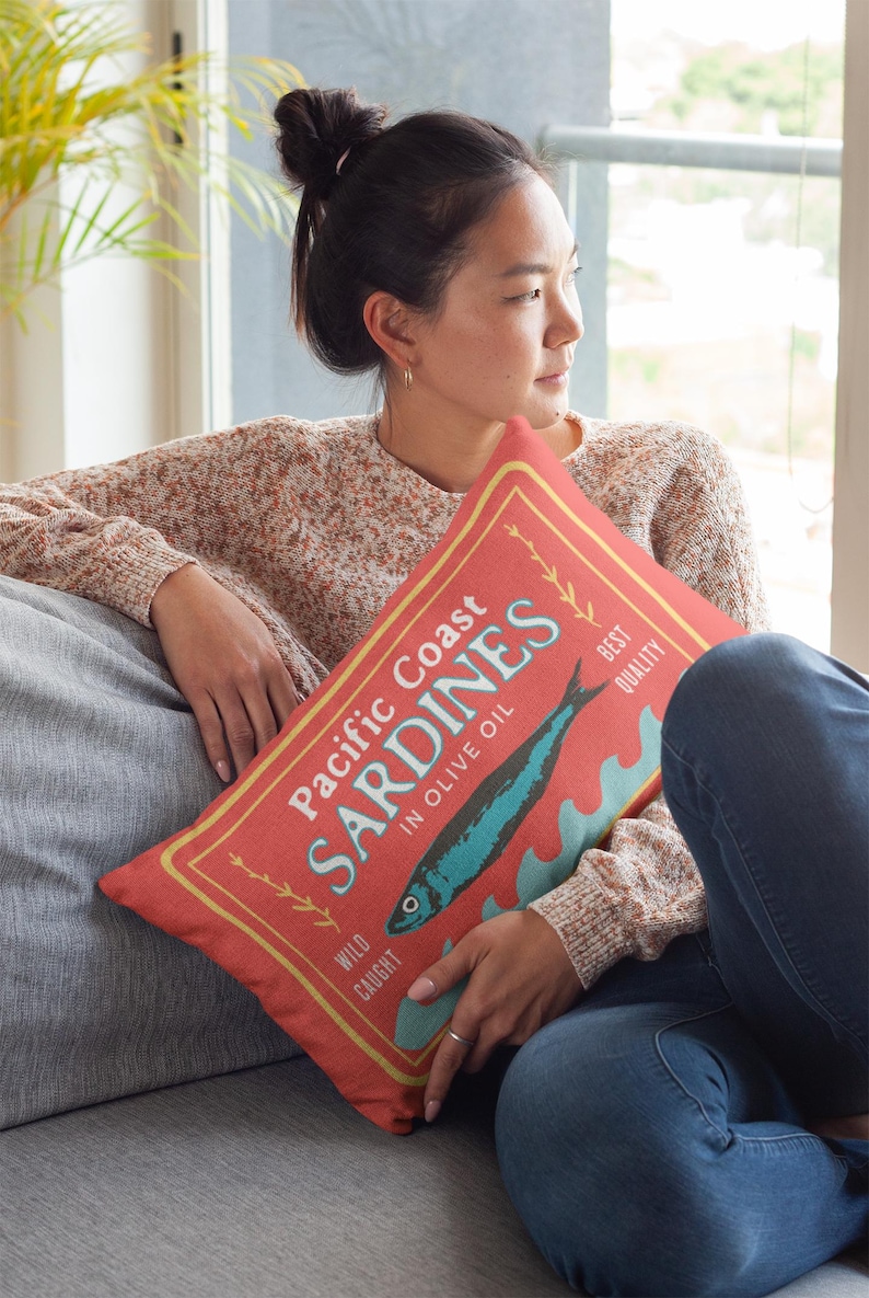 May include: Red decorative pillow with a vintage-style sardine can label design. The label reads "Pacific Coast Sardines in Olive Oil" with a blue fish illustration and text in a yellow border. The pillow is rectangular.
