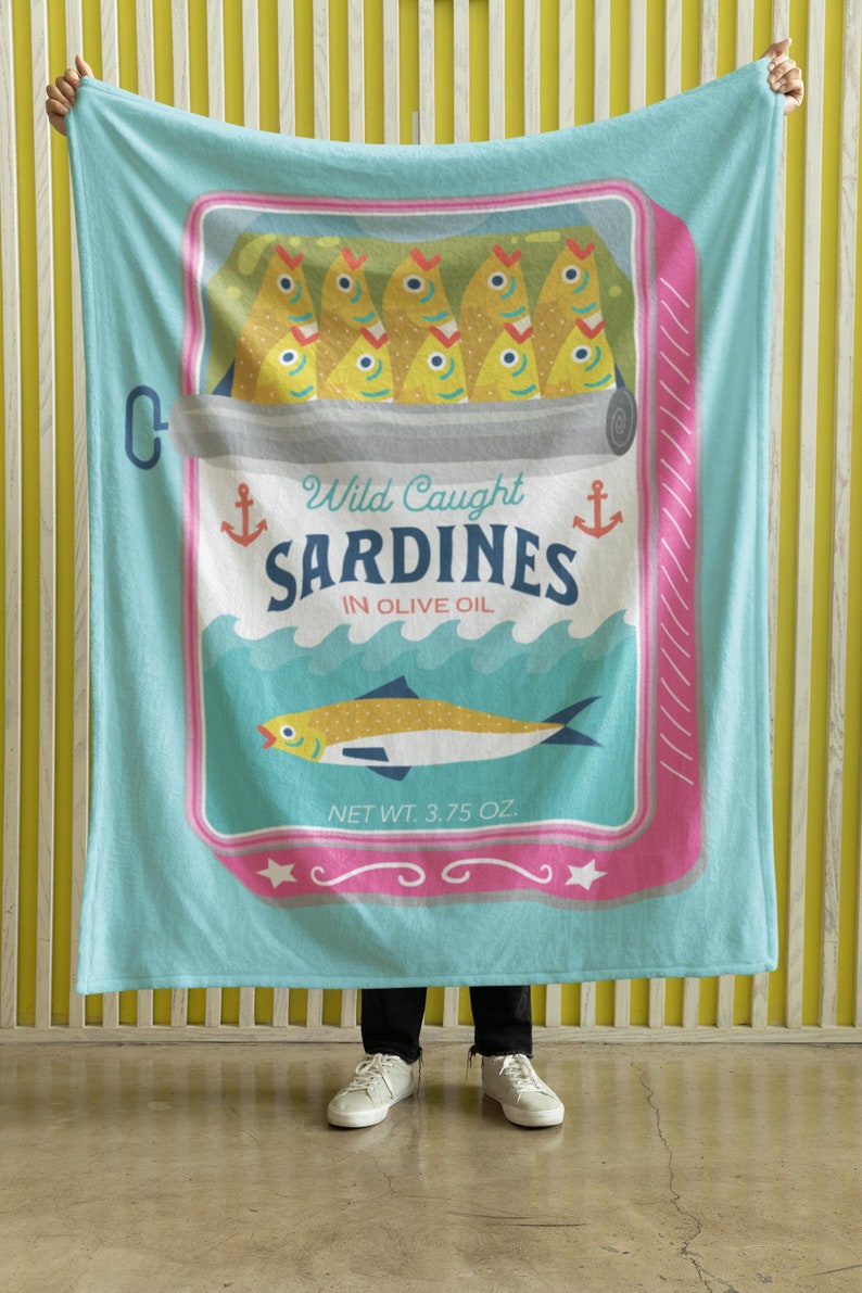 May include: A turquoise blue throw blanket with a vintage-style illustration of a can of sardines. The can has a pink and white border and says "Wild Caught Sardines in Olive Oil" with an anchor on each side. The can also has a picture of a sardine swimming in the ocean. The net weight is 3.75 oz.