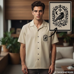 May include: A beige short-sleeve button-down shirt featuring a black graphic print on the chest pocket. The design depicts a raven perched on a branch, surrounded by roses, a crescent moon, a cross, and stars within an ornate oval frame.