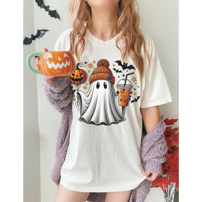 Puede incluir: Camiseta blanca con un fantasma de dibujos animados que lleva un sombrero y sostiene una taza de caf&eacute;. El fantasma est&aacute; rodeado de estrellas y murci&eacute;lagos. El texto "Boo!" est&aacute; impreso en la camiseta.