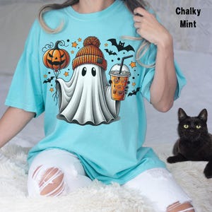 Puede incluir: Una camiseta azul turquesa con un fantasma de dibujos animados que lleva un gorro y sostiene una calabaza y una taza de caf&eacute;. El fantasma est&aacute; rodeado de estrellas y murci&eacute;lagos. El texto "Chalky Mint" est&aacute; en la esquina superior derecha de la imagen.