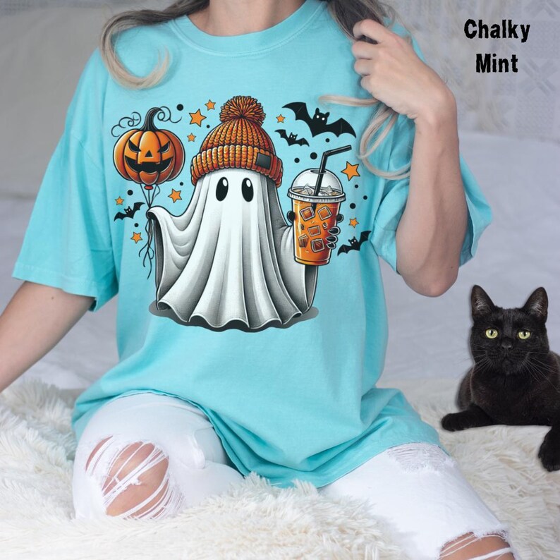 Puede incluir: Una camiseta azul turquesa con un fantasma de dibujos animados que lleva un gorro y sostiene una calabaza y una taza de caf&eacute;. El fantasma est&aacute; rodeado de estrellas y murci&eacute;lagos. El texto "Chalky Mint" est&aacute; en la esquina superior derecha de la imagen.