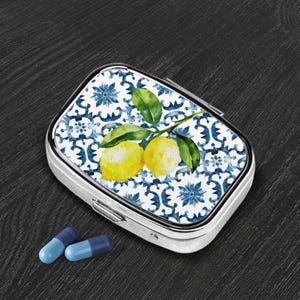May include: A silver pill box with a hinged lid, featuring a watercolor design of lemons and leaves on a blue and white patterned background. Two blue and light blue capsules are next to the box.