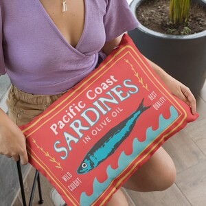 May include: A rectangular red pillow with the words "Pacific Coast Sardines in Olive Oil" in white and blue. A blue fish illustration is centered, with blue waves below. The pillow has a vintage, retro design.