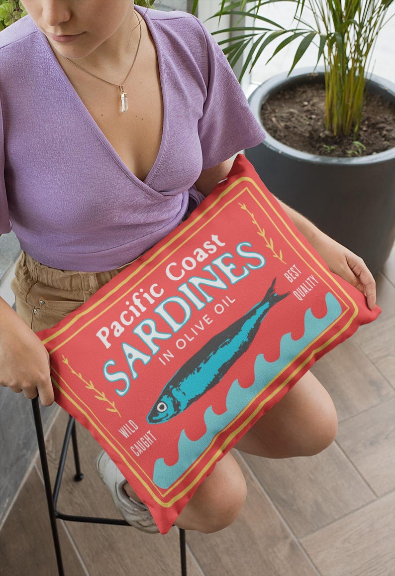 May include: A rectangular red pillow with the words "Pacific Coast Sardines in Olive Oil" in white and blue. A blue fish illustration is centered, with blue waves below. The pillow has a vintage, retro design.
