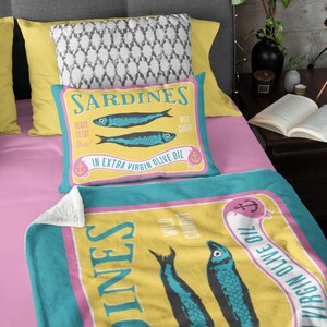 Puede incluir: Un juego de coj&iacute;n y manta con un dise&ntilde;o de sardinas. El coj&iacute;n y la manta tienen un fondo amarillo con detalles en azul y rosa. El dise&ntilde;o incluye dos sardinas azules y el texto "Sardines" y "In Extra Virgin Olive Oil."
