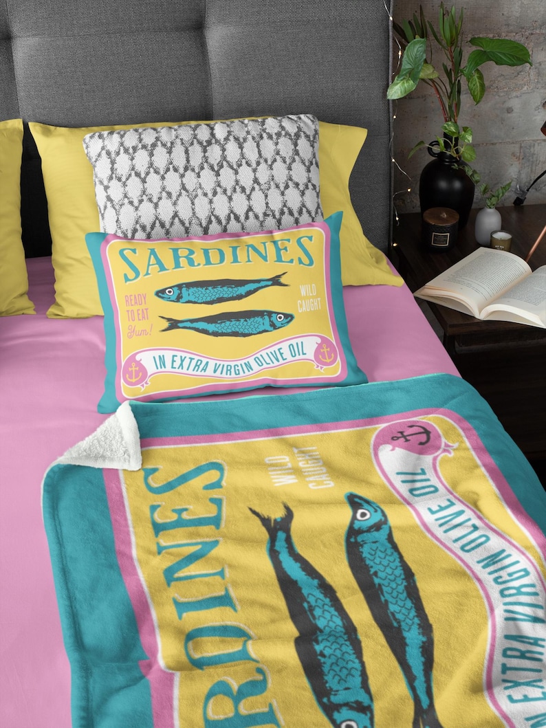 Puede incluir: Un juego de coj&iacute;n y manta con un dise&ntilde;o de sardinas. El coj&iacute;n y la manta tienen un fondo amarillo con detalles en azul y rosa. El dise&ntilde;o incluye dos sardinas azules y el texto "Sardines" y "In Extra Virgin Olive Oil."
