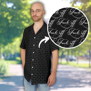 Puede incluir: Camisa negra de manga corta con un patrón repetido de las palabras "Fuck off" en escritura cursiva. La camisa tiene una botonadura y un cuello clásico. El fondo es una escena exterior borrosa.