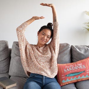 May include: A woman wearing a patterned sweater and blue jeans sits on a gray sofa. A decorative pillow with a sardine design and text is on the sofa. A book rests on the sofa, and a plant is in the background.