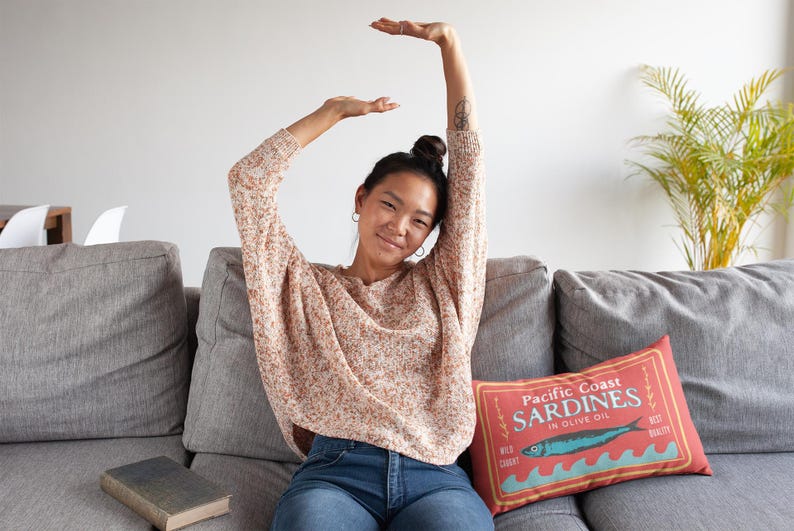 May include: A woman wearing a patterned sweater and blue jeans sits on a gray sofa. A decorative pillow with a sardine design and text is on the sofa. A book rests on the sofa, and a plant is in the background.