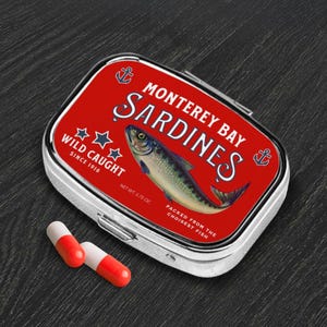 May include: A silver pill case with a red lid featuring the text "Monterey Bay Sardines" and a fish illustration. The case also has the text "Wild Caught Since 1916" and two red and white capsules are next to it.