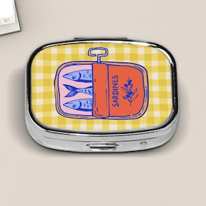 Peut inclure: Une bo&icirc;te &agrave; pilules argent&eacute;e avec un motif vichy jaune. Le couvercle pr&eacute;sente une illustration de dessin anim&eacute; d'une bo&icirc;te de sardines avec des poissons bleus, une &eacute;tiquette orange qui dit "Sardines" et une branche de feuilles.