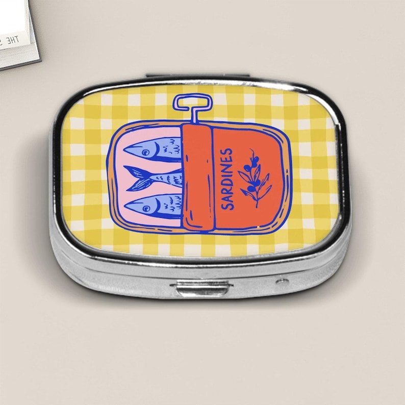 Peut inclure: Une bo&icirc;te &agrave; pilules argent&eacute;e avec un motif vichy jaune. Le couvercle pr&eacute;sente une illustration de dessin anim&eacute; d'une bo&icirc;te de sardines avec des poissons bleus, une &eacute;tiquette orange qui dit "Sardines" et une branche de feuilles.
