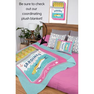 May include: A pink and blue plush blanket featuring a design of a sardine can. The can has a blue and white label with the text "Wild Caught Sardines in Olive Oil" and a net weight of 3.75 oz. The blanket is on a bed with two pillows with a geometric pattern.