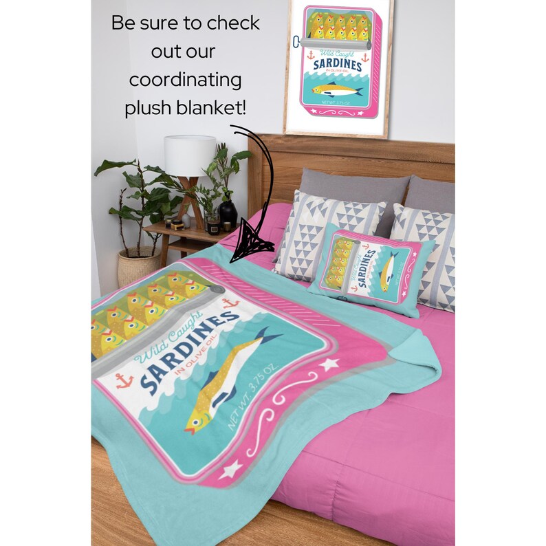 May include: A pink and blue plush blanket featuring a design of a sardine can. The can has a blue and white label with the text "Wild Caught Sardines in Olive Oil" and a net weight of 3.75 oz. The blanket is on a bed with two pillows with a geometric pattern.