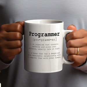 Puede incluir: Taza de cerámica blanca con un gráfico negro que define la palabra "Programador" e incluye una definición humorística. La taza está sostenida por la mano de una persona.