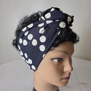 Pode incluir: Uma faixa de cabelo de tecido azul-marinho escuro com um padrão de bolinhas brancas. A faixa é amarrada com um nó no centro superior e exibida em uma cabeça de manequim com cabelo escuro e cacheado, cobrindo a testa e as laterais da cabeça.
