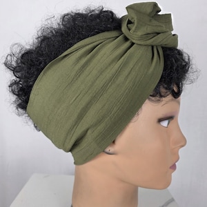 Peut inclure: Un bandeau en tissu vert olive présentant un aspect texturé et froissé avec un détail noué sur le dessus. Le bandeau est enroulé autour d'une tête de mannequin aux cheveux courts, foncés et bouclés, mettant en valeur le drapé du tissu.