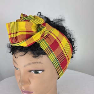 Peut inclure: Un bandeau en tissu arborant un motif à carreaux vif dans des tons de jaune, rouge et vert. Le bandeau est noué sur le dessus et présenté sur une tête de mannequin aux cheveux noirs courts et bouclés, mettant en valeur la texture textile.