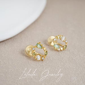 May include: A pair of gold-toned stud earrings. Each earring features a circular design adorned with green, white, and clear gemstones. The earrings have a post backing. The text "Lafaifa Jewelry" is in cursive script at the bottom.