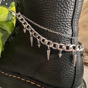 May include: Close-up of a black leather boot with a silver chain ankle bracelet. The chain features large links and dangling silver spikes. The boot has a thick sole and yellow stitching.