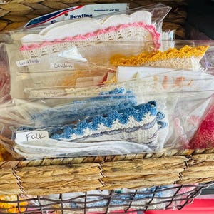 May include: A wicker basket filled with plastic bags containing various types of lace and fabric. The bags are labeled with the words "Folk", "Lace Doily", "Crochet", and "Pallison".
