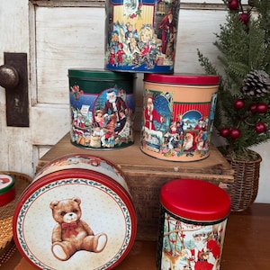 May include: A collection of vintage Christmas tins in various sizes and shapes. The tins feature festive illustrations of Santa Claus, children, and winter scenes. One tin has a teddy bear design. The tins are red, green, blue, and orange.