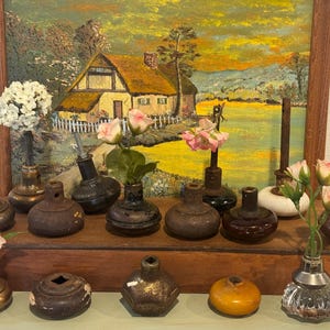 May include: A collection of antique glass and metal bud vases, each holding a single flower or small bouquet. The vases are displayed on a wooden shelf in front of a framed landscape painting. The vases vary in size, shape, and color.