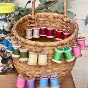 May include: A wicker basket filled with colorful wooden spool earrings. The spools are painted in various shades of pink, red, green, blue, and yellow. The earrings are displayed in pairs, with each pair featuring a different color.
