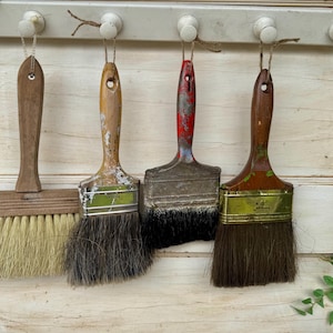 May include: Four vintage paint brushes with wooden handles hanging from a white peg rack. The brushes feature varying bristle colors including light tan, dark gray, black, and brown, with metal ferrules showing signs of wear and paint residue.