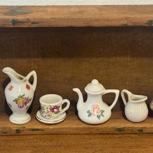 May include: A collection of miniature porcelain tea set pieces, including a teapot, creamer, sugar bowl, and teacup with saucer. The pieces are white with floral and gold accents, displayed on a wooden shelf.