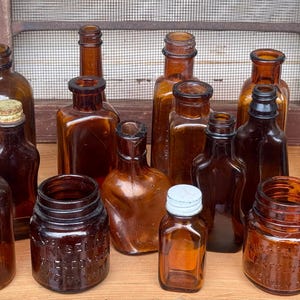May include: A collection of fifteen amber-colored glass bottles of various shapes and sizes. The assortment includes square, round, and rectangular bottles, some with narrow necks and others with wide openings. One bottle features a white metal cap.