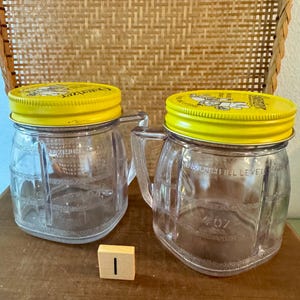 May include: Two clear glass jars with yellow lids and handles. The jars have a 4 oz fill level marking. The lids have text and graphics. A wooden block with the number 1 is in front of the jars. The jars are on a wooden surface.