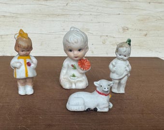 Pode incluir: Uma coleção de figuras de porcelana vintage. O conjunto inclui três pequenas figuras de bonecas e uma figura de cordeiro. As bonecas estão vestidas de branco e têm traços faciais pintados. O cordeiro é branco com uma coleira vermelha. As figuras são exibidas em uma superfície de madeira.