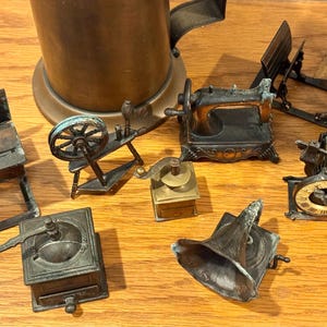 May include: A collection of miniature antique metal objects, including a coffee grinder, sewing machine, spinning wheel, and gramophone. The items are in shades of bronze and copper, arranged on a wooden surface.