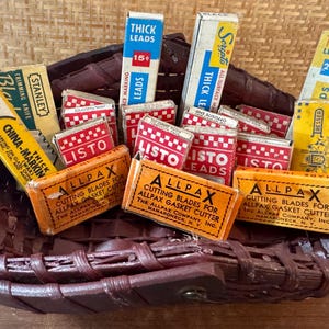 May include: A collection of vintage boxes of leads, blades, and a trimming knife, arranged in a dark brown woven basket. The boxes feature text like "LISTO LEADS" and "ALLPAX CUTTING BLADES".