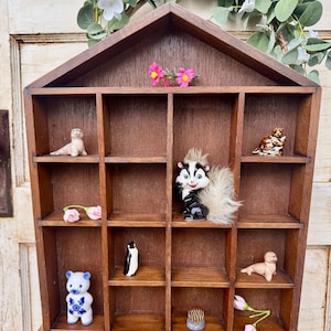 May include: A wooden display case with a house-shaped roof, featuring 12 compartments. The case is filled with various small figurines, including a penguin, a bear, a skunk, and several seals. The case is a great way to display small collectibles.