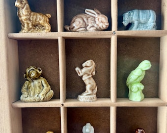 May include: A wooden display case with nine ceramic figurines of various animals, including a dog, rabbit, horse, monkey, parrot, and a few others. The figurines are arranged in a grid pattern.