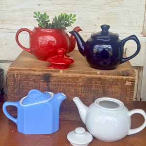 May include: Four ceramic teapots in red, navy blue, light blue, and white. The red teapot holds greenery. The teapots are displayed on a wooden box with text that reads "BARBERS SHAVING SOAP".