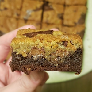 May include: A close-up of a brownie with a layer of cookie dough on top. The brownie is a dark brown color and the cookie dough is a light brown color. There are chocolate chips visible in the cookie dough.