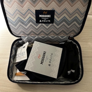 May include: Open travel kit with a patterned interior and the "Missoni Exclusively for Delta" logo. The kit contains earplugs, socks, and other travel accessories. The kit is black with a zipper closure.