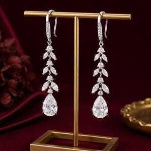 May include: A pair of silver-toned drop earrings featuring a vertical arrangement of marquise-cut and round-cut clear stones, terminating in a large pear-shaped stone. The earrings are suspended from a gold-toned T-shaped display stand.