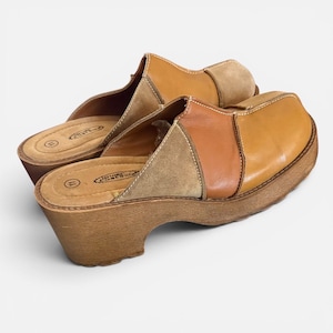 May include: A pair of slip-on clogs featuring a multi-tonal brown leather and suede upper with a square toe. The shoes have a thick, textured wood-look platform heel and a cushioned footbed with the text Lower Eastside and the number 11 printed.