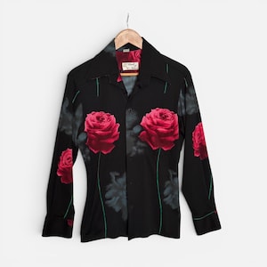May include: A black long-sleeve button-down shirt featuring a print of large red roses with green stems. The shirt has a wide collar and a label inside the neck that reads Touch of Class. The fabric displays a dark floral pattern throughout.