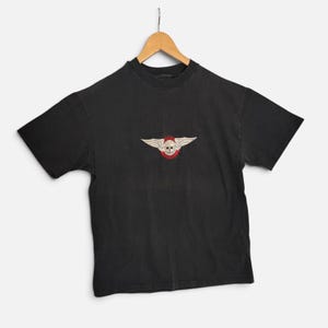 May include: A black short-sleeve t-shirt featuring a central graphic of a skull with white wings and a red circular border. The design includes the text "FAT BOY" above the skull and "MOTORCYCLES" below it, printed on a standard crew neck shirt.