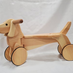 May include: A wooden toy dachshund with a brown stain finish. The toy has a handle on the head and two wheels on the back. The toy is made of natural wood and is suitable for children.
